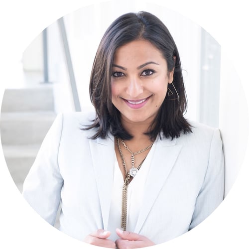 Dee Patel smiles at the camera, wearing a white blazer and standing near a set of stairs. 