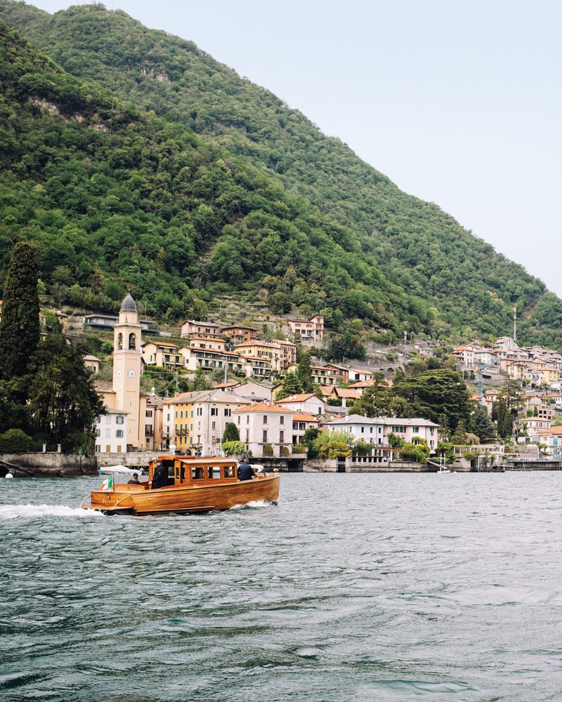 A wooden boat cruises on Lake Como, backdropped by a village spread out along the bottom of a forested hill.