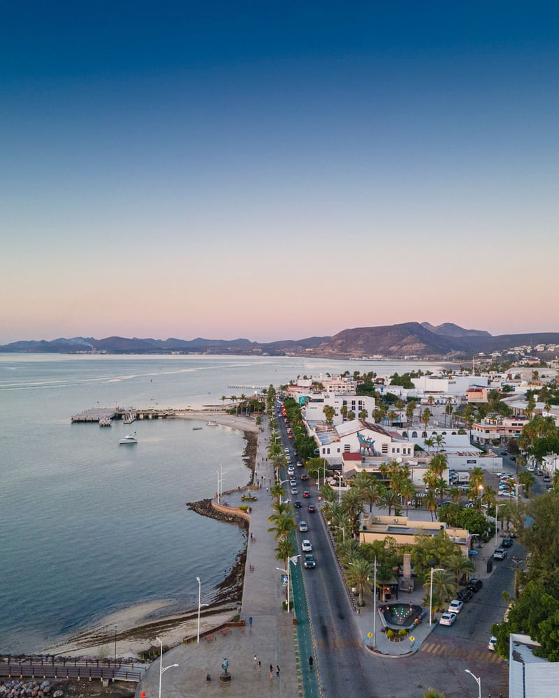 A waterfront road and neighborhood seen from above in La Paz, Mexico, at sunset.