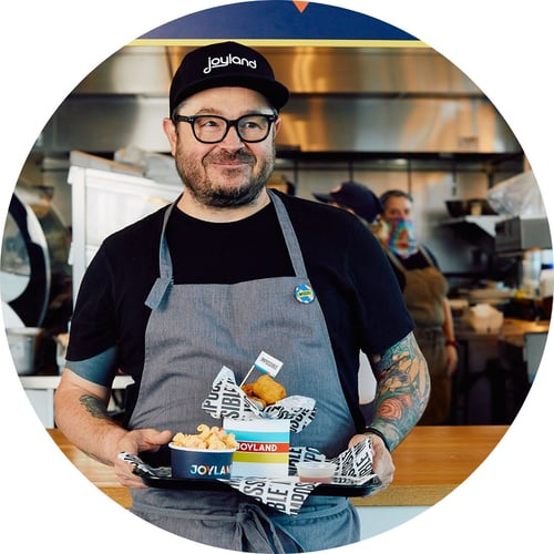 Chef Sean Brock, wearing black clothes and a gray apron, holds a tray of food from Joyland while grinning.