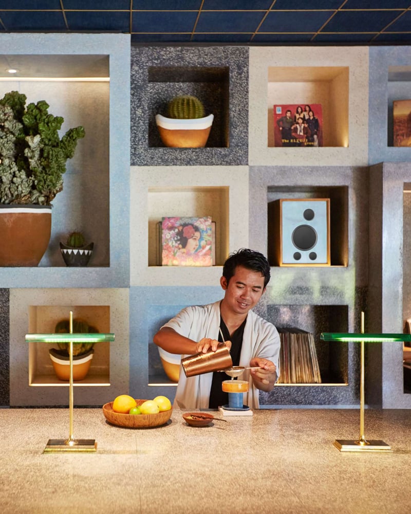 A bartender at Potato Head Suites pours a cocktail in front of a wall with modular shelves displaying cacti, records, and a speaker.