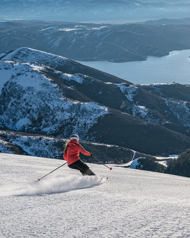 A skier in a red jacket heads downhill in Deer Valley, Utah, with a river visible in the distance.