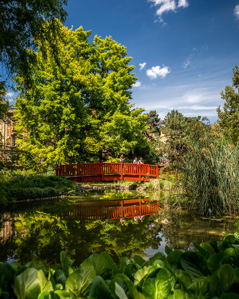 Park-goers cross a red bridge over a lake surrounded by greenery in Zagreb.